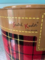 Close up side view of the cooler showing the Skotch Kooler logo, 4 gal capacity, and red tartan plaid pattern with yellow stitching details.