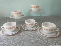 Five white ceramic cups and saucers displayed on a table, white background with light green tiled wall, showing floral patterns around rims