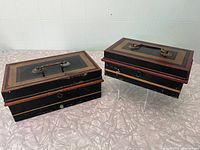 Two vintage metal cash boxes with removable trays, shows top view with handles and decorative trim in black and dark red-orange, placed on a silver patterned surface.