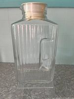 Front view of clear glass juice pitcher showing vertical ribbed pattern, rectangular shape, integrated handle and beige plastic top.