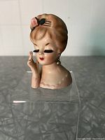 Front view showing stylized woman's face, closed eyes with long eyelashes, pink flower in hair, and one hand near face with painted nails