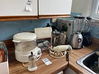 Wide view of kitchen counter showing multiple appliances including white Black and Decker steamer, hand mixer box, AeroGarden box, Bamix immersion blender, French press, toaster, and two blenders.