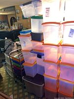 Multiple stacked plastic bins with orange and blue lids, transparent bodies, situated indoors.