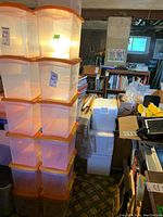 Stack of seven clear plastic storage bins with orange lids, arranged in two columns, shown in a basement storage area.