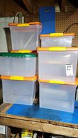 Stack of clear plastic storage bins with orange lids featuring yellow latches and one with green lid, showing size variation.