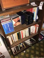 Wooden bookcase shelf containing assorted CDs in jewel cases stacked horizontally, and some hardcover and paperback books on adjacent shelves.