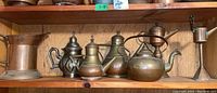 Shelf display of seven brass and copper metal items including kettle, teapots, a pitcher, and oil lamps with visible age patina