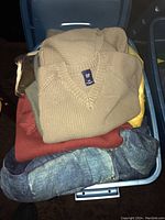 Bin filled with multiple men's clothing items including sweaters and shirts, folded and visible from above.