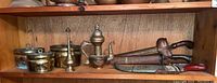 Shelf display of three brass pots, a vintage-style perfume bottle, a brass teapot, and three weapons including the kris sword with wooden and metal sheath and other sheathed blades.