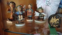 Five ceramic Hummel figurines in various poses, colors, and outfits, displayed on a shelf.