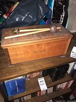 Rectangular wooden box with three slits on top and two mallets resting on it, placed on a shelf among books and bags.