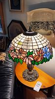 Full view of the lamp including stained glass style floral shade and metal base on wooden table next to dark furniture and framed artwork.