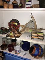 Shelf displaying the red mask, owl figurine on driftwood, trinket box, and colorful decorative bowl with mugs below