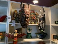 Shelf display of four decorative masks and one figurine arranged together with a small green clock visible below.