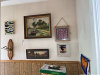 Wall with multiple framed artworks including main signed oil painting and smaller prints along with wool wall hangings