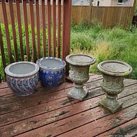 Four plant pots outdoors on a wooden deck: two ceramic pots with floral patterns and two plastic urn-shaped pots with ornate detailing.