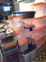 Stacked clear plastic storage bins with orange lids, topped with bins with blue and black lids, and a large gray bin at the bottom.