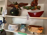 Photo of two white shelves holding pottery and figurines including black vase, pottery bowls, and frog wood figurines