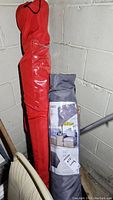 Two patio umbrellas in their respective carrying cases, red and dark grey, against a basement concrete wall.