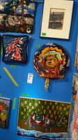 Five wall art pieces on display on a blue wall, including yarn art, hook rugs depicting fish and an elephant, and a framed print.