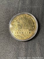 Close up front image of 1oz History of Gold coin inside clear plastic protective cover showing detailed relief with buildings and graphical elements.