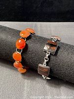Photo showing two chunky bracelets on black display stand; one with orange-red gemstones, the other with dark wood-like insets