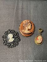 Three Victorian style cameo pendants displayed on a dark fabric background, showing size and detail differences.