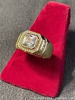 Ring displayed on a red ring stand on grey fabric showcasing front view with large clear Asscher cut stone and surrounding smaller stones.