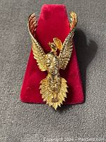 Close-up image of gold-tone flying phoenix brooch displayed on red velvet stand showing detailed wings and tail feathers.