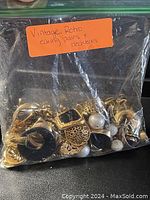 Photo of a plastic bag containing numerous vintage and retro style earrings and necklaces mostly gold-tone with some black and pearl accents.
