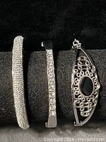 Front view of three silver tone bracelets laid side by side showing rhinestone designs and black gemstone bracelet.
