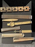 Photo of seven metal tie clips in various designs and finishes displayed on black backing.