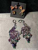 Photo showing both pairs of earrings on a flat surface: iridescent mesh bow with square iridescent studs on a black holder, and large peacock-themed drop earrings with colorful bead dangles.