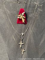 Photo showing three necklaces laid flat with silver-tone chain having a bird charm and rhinestone cross, and a gold-tone cross fish pendant on a thin chain alongside a gold-tone angel brooch on red velvet stand.