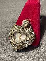 Heart shaped rhinestone-encrusted pendant with embedded clock displayed on red velvet ring holder, showcasing front side and clock face in detail.