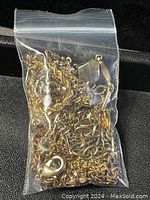 Small clear resealable plastic bag containing a tangled mass of metal chains in gold, silver, and bronze colors. Various chain styles and clasps are visible through the bag.
