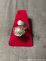 Front top view of silver adjustable ring on red stand showing floral and ornate designs on spoon handle band