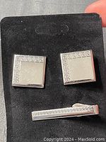 Photo showing two square silver tone cufflinks and one matching tie clip on black velvet background. The Greek key pattern borders one edge of each piece.