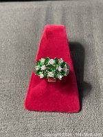 Close-up photo of the front of the ring showing green emerald stones and white diamond stones set in a flower pattern on a silver-tone band.