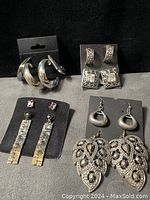 Photo showing all 7 pairs of earrings displayed on black earring holders, exhibiting diverse silver tone styles including hoops, filigree, geometric drops, and ornate leaf designs with rhinestones.