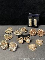 Full view of all 11 pairs of vintage pearl-themed earrings arranged on black background showing various designs including clusters, bows, hearts, and leaves in cream, pink, and gold tones.