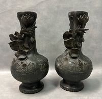 Pair of antique bronze vases photographed against a plain background, showing details of raised floral design on the bulbous body and neck, with signs of damage and wear.