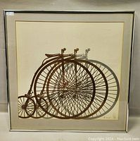 Full view of framed print showing a stylized tricycle bicycle design with large overlapping wheels in brown tones.