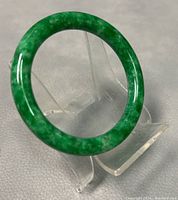 Green jade bangle displayed standing on clear plastic stand on gray textured surface.