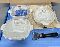 Three white square saucepans with clear glass lids and a black detachable handle in original packaging.