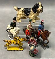Front view of vintage hand painted metal toys including a dog, three mounted soldiers, a drummer, and a yellow hunting dog, showing details and paint condition.