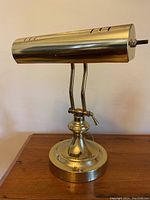 Front view of the brass adjustable banker’s desk lamp showing the cylindrical shade with ventilation slits and adjustable supports connected to the round base.