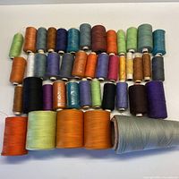 Photo showing multiple spools of serging thread in assorted colors laid out on a white surface, including greens, browns, blues, orange, black, and purple.
