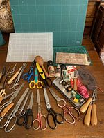 Full overview of sewing tools showing cutting mats, numerous scissors, spools, ink stamp letters, and rug hooking tools on wooden table.