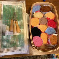 Photo of large latch hooking fabric piece with three latch hooking tools and a basket containing thirteen bundles of wool yarn.